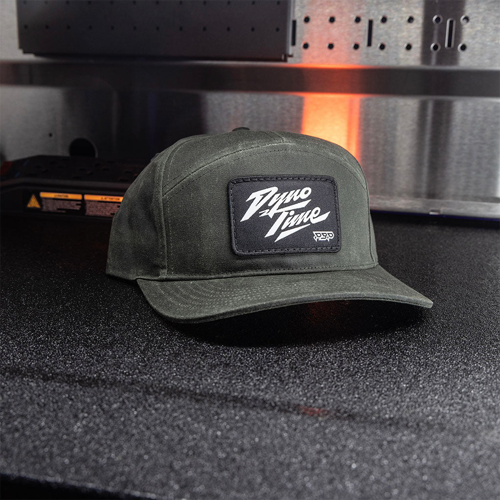 Dark olive green adjustable snapback hat with woven black patch with white dyno time text. 